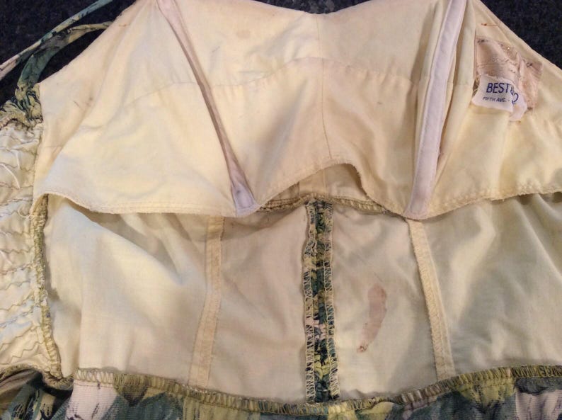 May include: The inside of a white dress with a green and yellow floral print. The dress has a built-in bra and a zipper in the back. The label says "Best 2" and "South Korea".