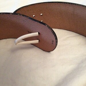 May include: A brown leather belt with a white buckle. The belt has a wide design and a classic style. The buckle is a simple, curved shape. The belt is likely for waist or hip wear.