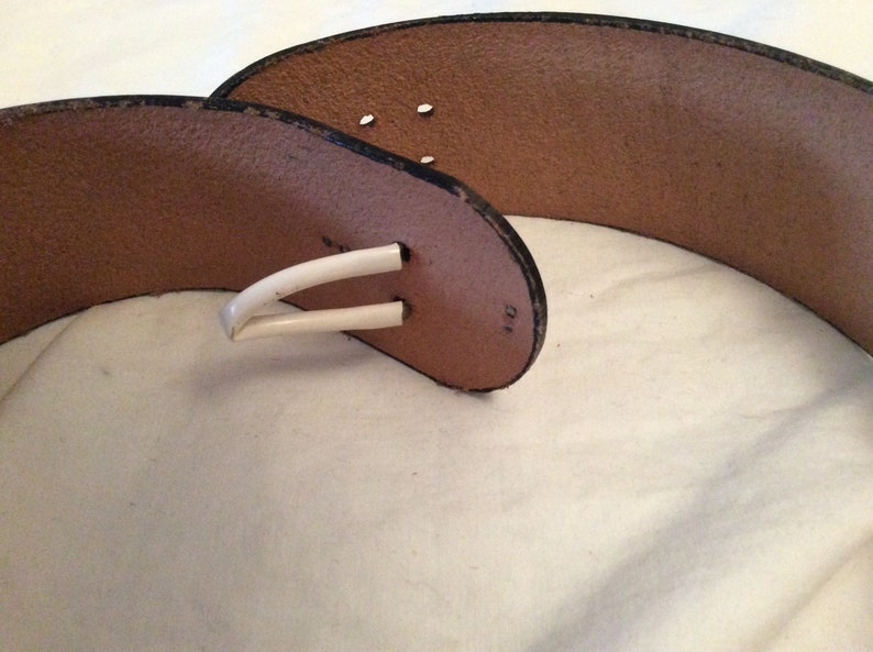 May include: A brown leather belt with a white buckle. The belt has a wide design and a classic style. The buckle is a simple, curved shape. The belt is likely for waist or hip wear.