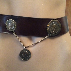 May include: A brown leather belt with three decorative coin medallions. The belt has a wide band with two large medallions on either side, connected by a gold chain to a third, smaller coin.