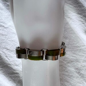 May include: A silver-toned bracelet with rectangular green and silver links. The bracelet is displayed on a white surface, showcasing its design and color contrast. The bracelet's links are approximately 0.75 inches long.