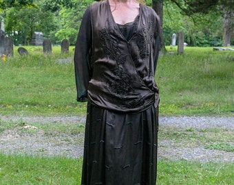 May include: A woman in a brown, beaded, silk dress stands in a grassy area with a cemetery in the background.