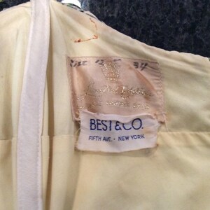 May include: A white label with the text "BEST & CO. FIFTH AVE. - NEW YORK" on a yellow background. Above the label is a pink label with the text "Lord & Taylor" and a crown.