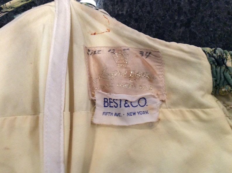 May include: A white label with the text "BEST & CO. FIFTH AVE. - NEW YORK" on a yellow background. Above the label is a pink label with the text "Lord & Taylor" and a crown.