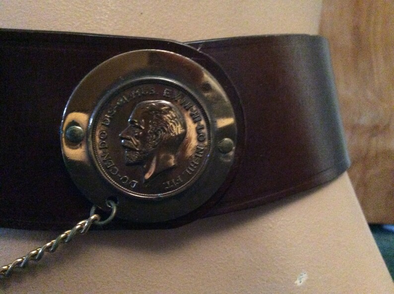 May include: A wide, dark brown leather belt with a large, circular metal buckle. The buckle features a detailed relief of a man's profile and text. A decorative chain hangs from the buckle.