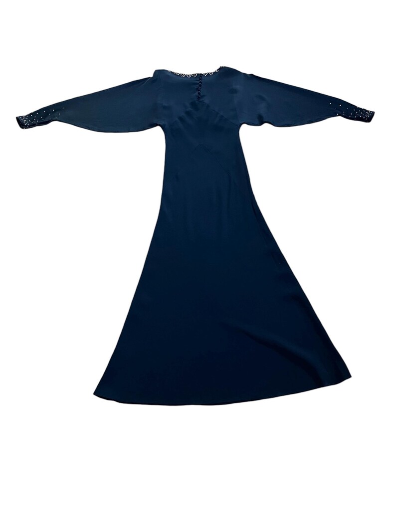 May include: A long, dark blue dress with a V-neckline and long sleeves. The sleeves have a row of small, white embellishments at the cuffs.