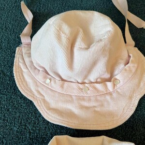 K&ouml;nnte beinhalten: Ein hellrosa Sonnenhut aus Cord mit breiter Krempe und rundem Oberteil. Der Hut hat ein Kinnband und wird mit drei kleinen Kn&ouml;pfen befestigt. Der Hut ist f&uuml;r den Sonnenschutz konzipiert.