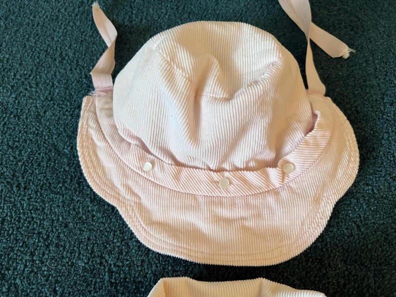 K&ouml;nnte beinhalten: Ein hellrosa Sonnenhut aus Cord mit breiter Krempe und rundem Oberteil. Der Hut hat ein Kinnband und wird mit drei kleinen Kn&ouml;pfen befestigt. Der Hut ist f&uuml;r den Sonnenschutz konzipiert.