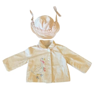 K&ouml;nnte beinhalten: Hellrosa Baby-Outfit-Set, bestehend aus einer Jacke und einer M&uuml;tze. Die Jacke hat eine Knopfleiste und ein gesticktes G&auml;nsemotiv. Die M&uuml;tze hat einen ger&uuml;schten Rand und Bindeb&auml;nder. Das Set ist aus einem weichen, hellfarbigen Stoff.