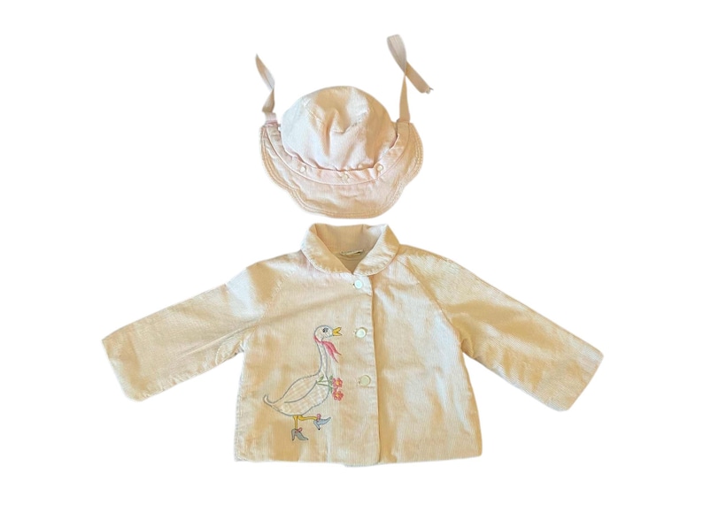 K&ouml;nnte beinhalten: Hellrosa Baby-Outfit-Set, bestehend aus einer Jacke und einer M&uuml;tze. Die Jacke hat eine Knopfleiste und ein gesticktes G&auml;nsemotiv. Die M&uuml;tze hat einen ger&uuml;schten Rand und Bindeb&auml;nder. Das Set ist aus einem weichen, hellfarbigen Stoff.