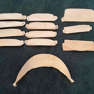 May include: A collection of beige fabric pieces, including several rectangular shapes with tabs, curved pieces, and a larger, arched component. The items are arranged on a dark green surface, likely for a craft or sewing project.