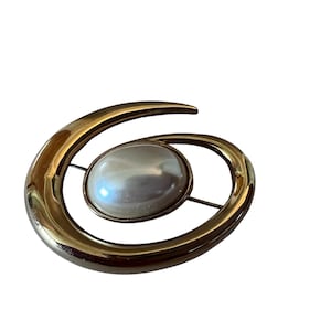 May include: A gold-tone metal brooch featuring a smooth, curved, crescent-like frame that wraps around a central, oval-shaped, white faux pearl cabochon. The polished metallic surface reflects light, and a thin pin mechanism is visible on the back.