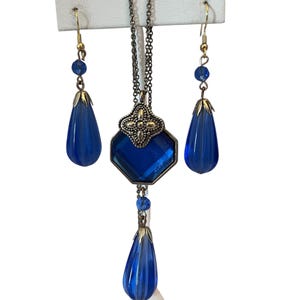 May include: A jewelry set consisting of a necklace and two matching earrings. The necklace features a dark metal chain with a central hexagonal blue glass pendant and a smaller dangling blue glass teardrop. The earrings have blue glass teardrops.