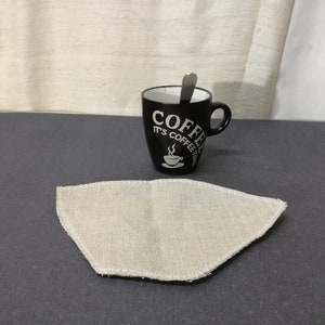 Op de afbeelding: Een wit katoenen koffiefilter ligt op een donkergrijs oppervlak naast een zwarte koffiemok met de tekst "Coffee It's Coffee Time" in wit.
