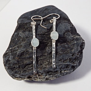 May include: Silver earrings with a hammered finish. Each earring has a vertical bar design with a light blue, oval stone. The earrings are displayed on a dark grey rock against a white backdrop.