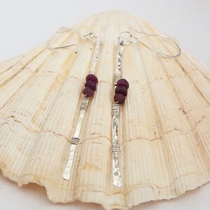 May include: Silver drop earrings with a hammered finish and three dark red gemstones. The earrings feature a simple, elegant design with wire-wrapped detailing. The earrings are displayed on a seashell.