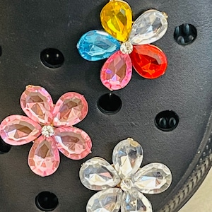 May include: Four flower-shaped charms with rhinestones in various colors, including pink, blue, yellow, and clear, attached to a black surface.