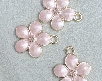May include: Three pink flower charms with gold-colored edges and loops. Each charm has five petals and a textured center. The charms are set against a light gray background.