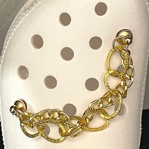 May include: A gold chain charm with two different link styles, attached to a white shoe with holes. The chain is made of two different styles of links, one is a simple chain link and the other is a larger oval link.