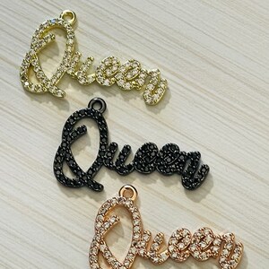 May include: Three charm pendants spelling out the word "Queen" in cursive script. Each pendant is made of metal and has a different finish: gold, black, and rose gold. Each pendant is adorned with clear rhinestones.