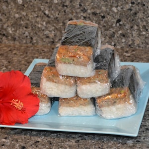 Może przedstawiać: Stos pięciu indywidualnie zapakowanych hawajskich Spam Musubi, popularne lokalne jedzenie na Hawajach, na jasnoniebieskim talerzu z czerwonym kwiatem hibiskusa.