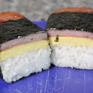 Może przedstawiać: Trzy hawajskie musubi ze Spam, każda z warstwą ryżu, cienką warstwą ż&oacute;łtego jajka, plastrkiem r&oacute;żowego Spam i warstwą wodorost&oacute;w.