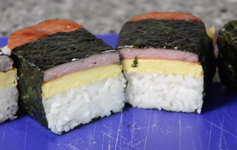 Może przedstawiać: Trzy hawajskie musubi ze Spam, każda z warstwą ryżu, cienką warstwą ż&oacute;łtego jajka, plastrkiem r&oacute;żowego Spam i warstwą wodorost&oacute;w.