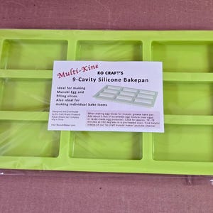 Może przedstawiać: Zielona silikonowa forma do pieczenia z dziewięcioma prostokątnymi wnękami. Forma jest oznaczona jako "Multi-KiTLE KO CRAFT'S 9-Cavity Silicone Bakepan". Jest idealna do robienia jaj Musubi i plastr&oacute;w nadzienia, a także indywidualnych wypiek&oacute;w.