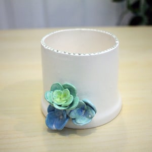 May include: White ceramic planter with a decorative beaded rim and three blue and green ceramic flowers.
