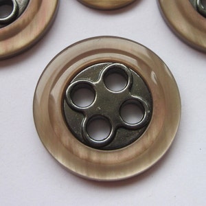 Könnte beinhalten: Ein großer, runder, brauner Knopf mit einem vierlöchrigen Metalldesign in der Mitte.