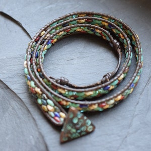 Könnte beinhalten: Ein mehrreihiges Wickelarmband mit braunem Lederband und bunten Glasperlen. Das Armband hat einen dreieckigen Anhänger mit einem dekorativen Design.