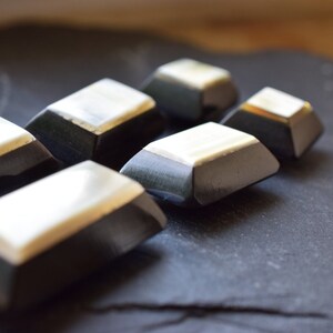 Könnte beinhalten: Nahaufnahme von sechs dunklen, geometrischen Knöpfen mit rechteckiger Form und einer hellen, möglicherweise elfenbeinfarbenen Oberseite. Die Knöpfe sind auf einer dunklen, strukturierten Oberfläche angeordnet und zeigen ihr einzigartiges Design.