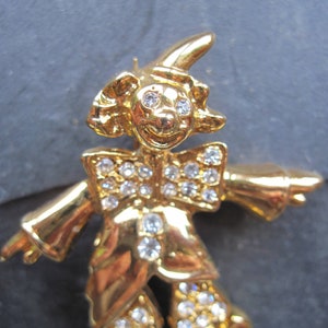 Könnte beinhalten: Eine goldfarbene Clown-Brosche mit einem roten Hut und weißen Strasssteinen. Der Clown trägt ein weißes Hemd mit einer Schleife und eine schwarze Hose.