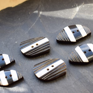 Könnte beinhalten: Sechs schwarz-weiß gestreifte Knöpfe mit jeweils zwei Löchern. Die Knöpfe haben eine strukturierte, holzähnliche Oberfläche.
