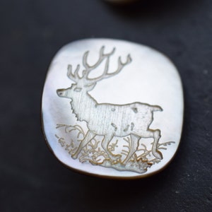 Könnte beinhalten: Ein weißer Perlmuttknopf mit einem detailliert gravierten Bild eines Hirsches, der in einem Grasfeld steht.