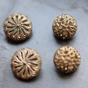 Könnte beinhalten: Vier verzierte goldfarbene Knöpfe mit komplizierten Designs. Die Knöpfe haben eine erhabene, strukturierte Oberfläche und eine runde Form.