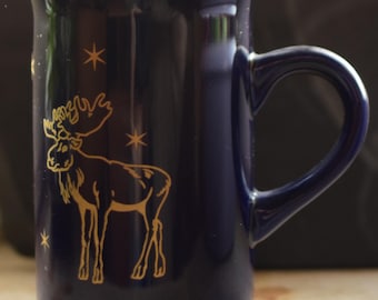 Könnte beinhalten: Eine blaue Keramiktasse mit einem goldenen Elch-Design und goldenen Sternen. Die Tasse hat einen großen Henkel.