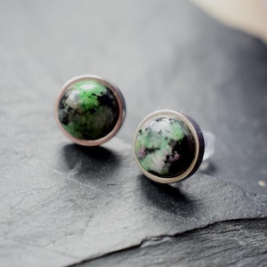 Könnte beinhalten: Ein Paar runde Ohrstecker. Jeder Ohrring hat einen gesprenkelten Edelstein in Grün, Schwarz und Rosa, der in einer silberfarbenen Lünette gefasst ist. Die Ohrringe werden auf einer dunklen, strukturierten Oberfläche präsentiert.