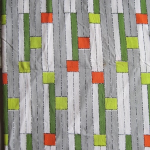 Könnte beinhalten: Ein weißer Stoff mit einem geometrischen Muster aus grünen, orangen und gelben Rechtecken. Der Stoff hat eine subtile Textur und ein sich wiederholendes Muster.