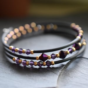Könnte beinhalten: Mehrstrang-Armband mit schwarzen Kordeln und lila-goldenen Perlen. Das Armband hat ein Wickeldesign mit drei Strängen. Die Perlen sind eine Mischung aus kleinen goldenen und facettierten lila Kristallen.