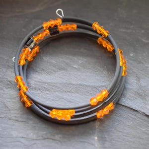 Könnte beinhalten: Ein mehrsträngiges Armband mit einem dunkelgrauen Band und orangefarbenen Perlen. Das Armband wickelt sich mehrmals um sich selbst und erzeugt einen geschichteten Effekt. Die orangefarbenen Perlen sind gleichmäßig über das Band verteilt.