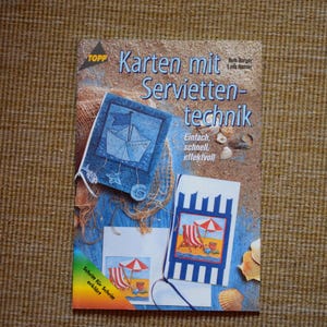 Könnte beinhalten: Ein Buch mit dem Titel "Karten mit Servietten-technik" von Ruth Burgos und Lenk Riebter. Der Buchumschlag zeigt ein nautisches Thema in Blau und Weiß mit einem Segelboot, Muscheln und einem Fischernetz. Der Text "Einfach, schnell, effektvoll" ist auf dem Umschlag gedruckt. Der Buchumschlag enthält auch den Text "Schritt für Schritt erklärt".