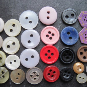 Könnte beinhalten: Eine Auswahl an Knöpfen in verschiedenen Farben und Größen, darunter weiß, rosa, rot, blau, braun und schwarz. Einige Knöpfe haben zwei Löcher, andere vier. Die Knöpfe sind aus verschiedenen Materialien hergestellt, darunter Kunststoff, Metall und Holz.