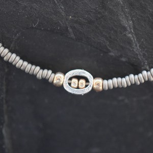 Könnte beinhalten: Ein silbernes Armband mit einem silbernen ovalen Anhänger mit drei kleineren silbernen Perlen. Das Armband besteht aus grauen Perlen.