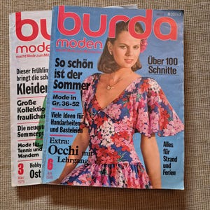 Könnte beinhalten: Zwei Burda Moden Zeitschriften mit blau-weißen Titelseiten. Das vordere Heft zeigt eine Person in einem Kleid mit rosa und roten Blumen auf blauem Grund, mit deutschem Text wie 'So schön ist der Sommer' und der Datumsangabe 'Juni 1978'.