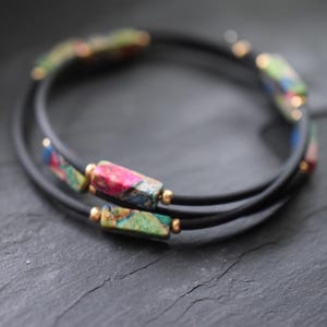 Könnte beinhalten: Wickelarmband mit schwarzen Schnüren und rechteckigen, farbigen Perlen. Die Perlen zeigen eine Mischung aus Rosa, Grün, Blau und Gold. Kleine goldfarbene Perlen akzentuieren das Design und schaffen ein stilvolles Accessoire.