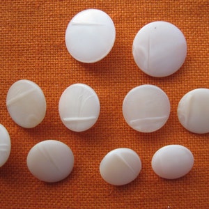 Könnte beinhalten: Zehn weiße runde Knöpfe mit einem erhabenen Design. Die Knöpfe sind in einem Halbkreis auf einem orangefarbenen Stoffhintergrund angeordnet.