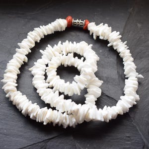 Könnte beinhalten: Weiße Muschelkette mit Spiralmuster. Die Kette besteht aus kleinen, unregelmäßigen Muschelteilen, die aneinandergereiht sind. Der Verschluss ist silberfarben mit roten Korallenakzenten. Die Kette wird auf einer dunkelgrauen Oberfläche präsentiert.