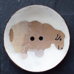 Könnte beinhalten: Eine weiße Keramikschale mit braunem Rand, auf der ein handgemaltes braunes Tier mit zwei Löchern in der Mitte abgebildet ist. Das Tier scheint ein Schaf oder eine Kuh zu sein.