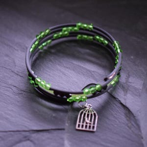 Könnte beinhalten: Ein dunkelgraues Wickelarmband mit grünen Perlen und einem silbernen Vogelkäfig-Anhänger. Das Armband hat ein Spiraldesign. Der Vogelkäfig-Anhänger ist detailliert und verleiht dem Accessoire eine einzigartige Note.